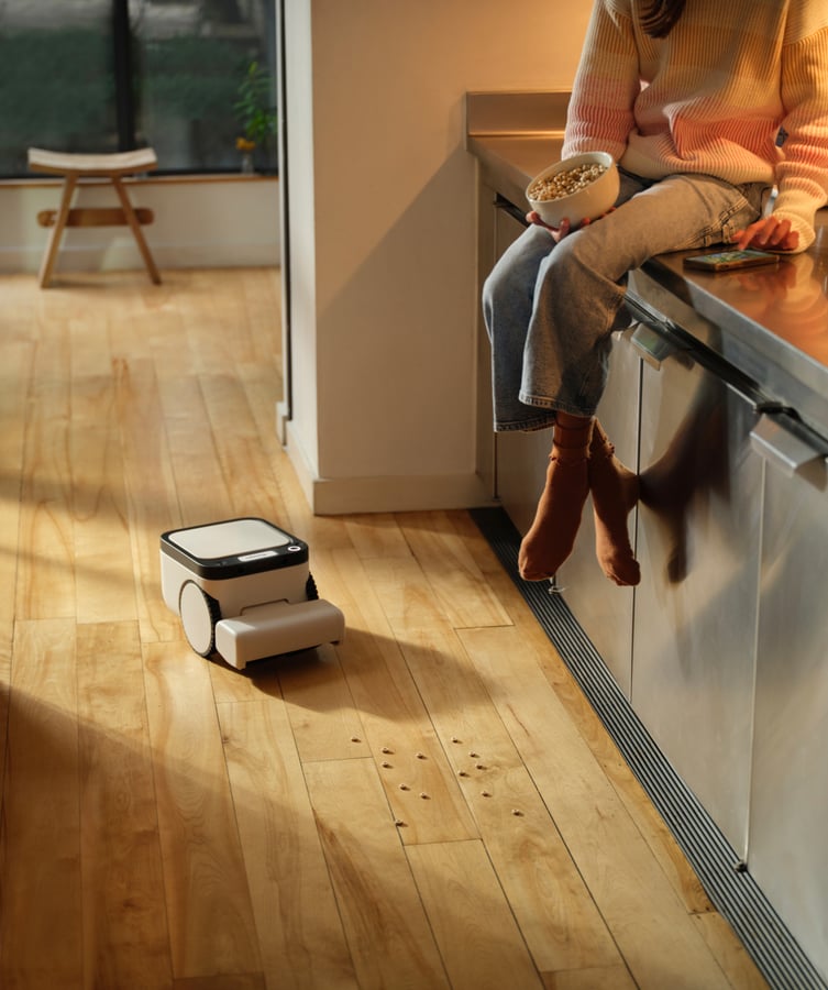 A child sitting on a kitchen counter with a bowl of cereal, spilled pieces on the floor and the Matic approaching them