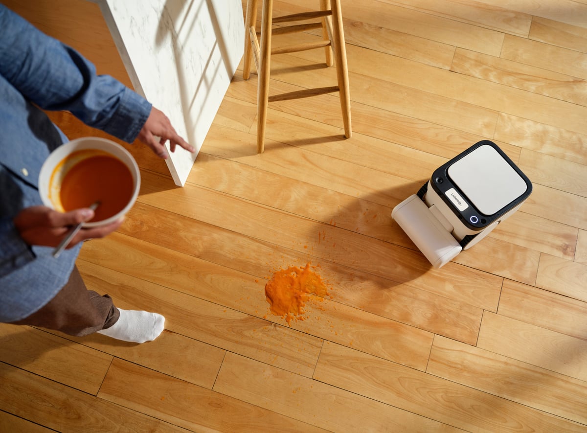 The Matic approaching a spilled bowl of soup on a hardwood floor, its display reading "Listening"