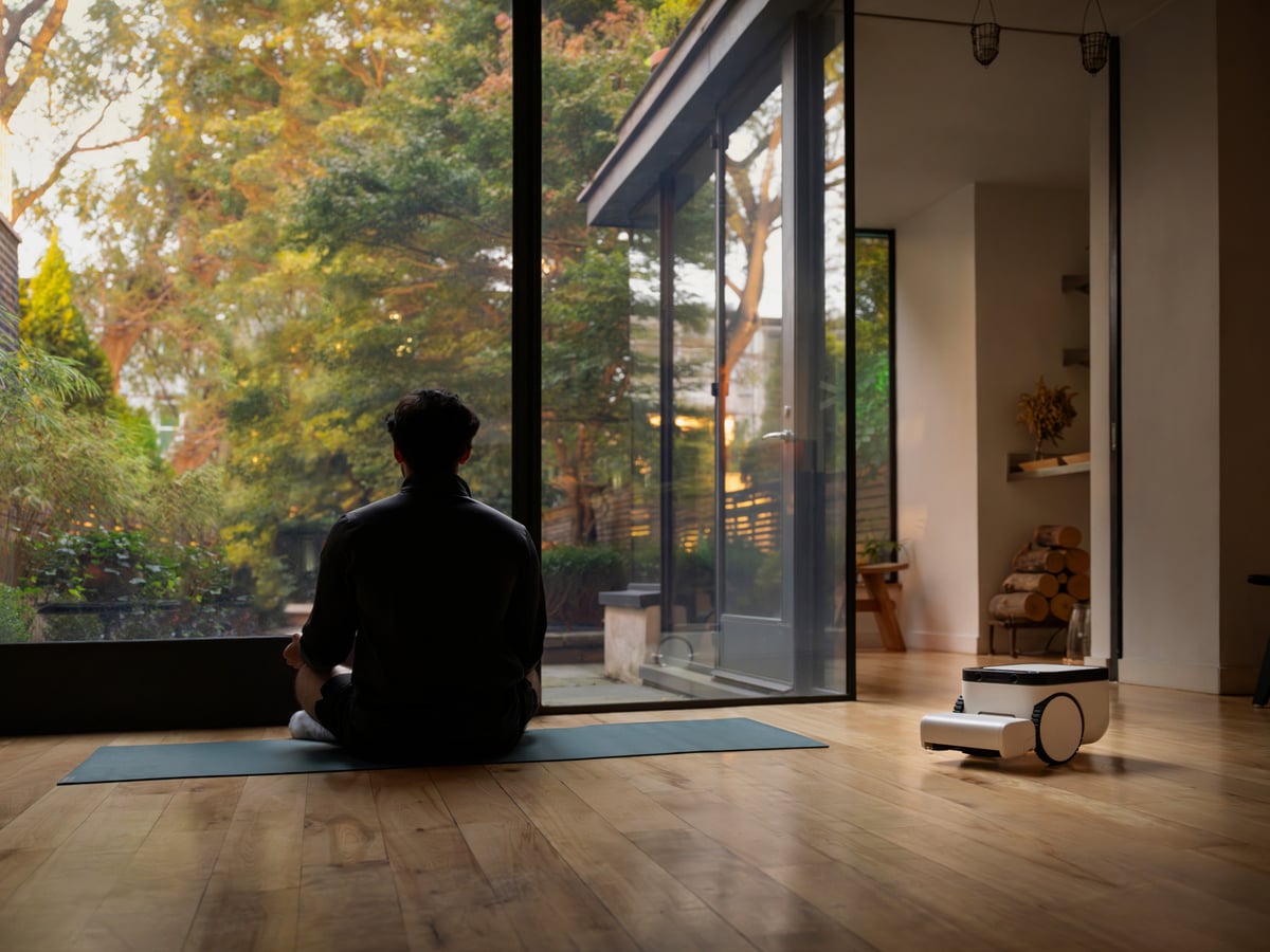 A person sitting cross-legged on a yoga mat by a garden window while the Matic works on the floor behind them
